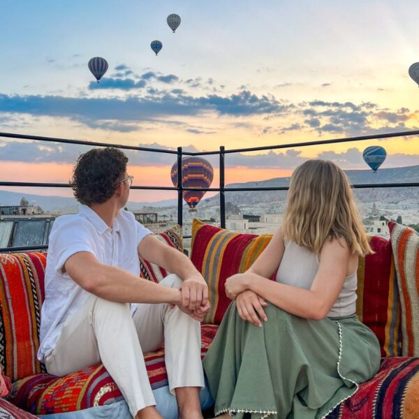 A young couple on the roof of a Cappadocia cave hotel with hot air balloons in the background.