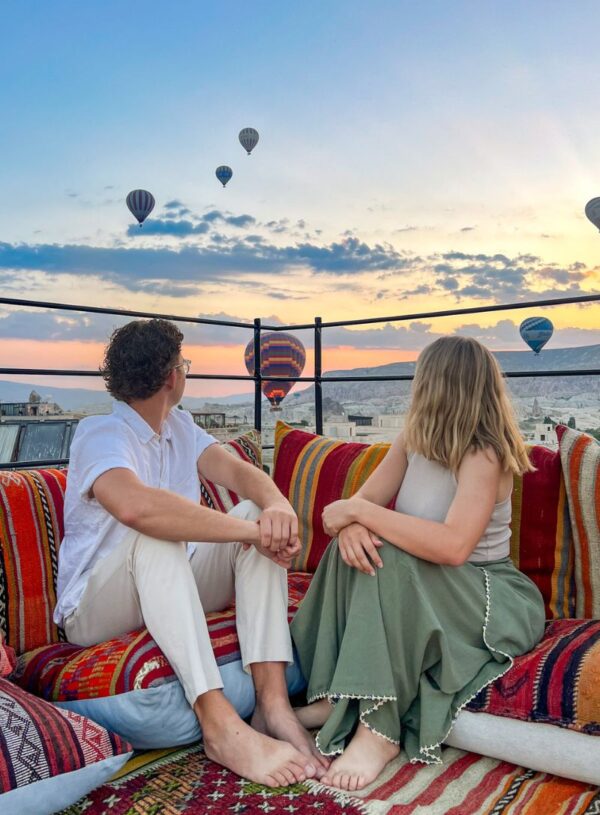 A young couple on the roof of a Cappadocia cave hotel with hot air balloons in the background.