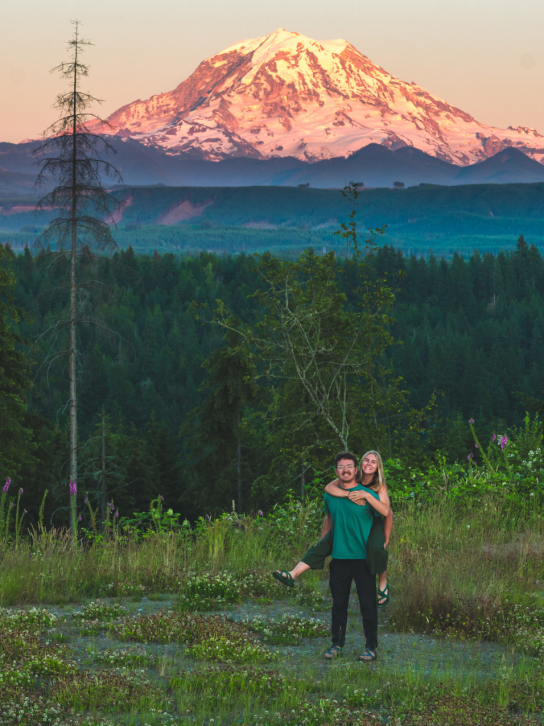 Couple smiling in a field at sunset with Mount Rainier glowing in the background, surrounded by wildflowers and forest, capturing a romantic moment on a Pacific Northwest road trip.