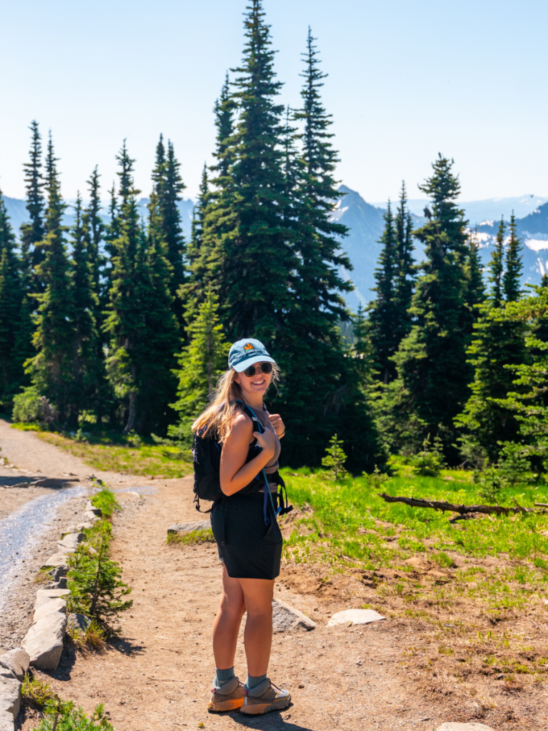Woman smiling on a sunny hiking trail surrounded by tall evergreens and mountain views in Mount Rainier National Park, enjoying a summer adventure on a Pacific Northwest road trip.