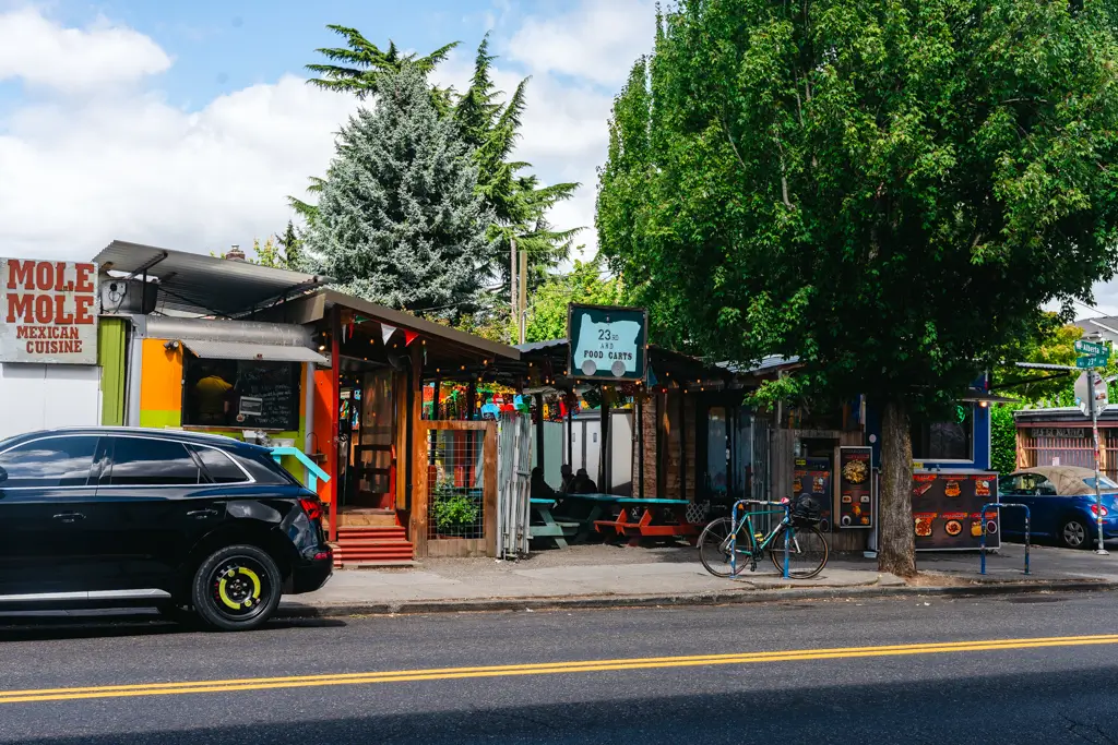 Street view of the 23rd Avenue Food Carts in Portland, Oregon, featuring colorful food stands, outdoor picnic tables, bicycles, and leafy trees.
