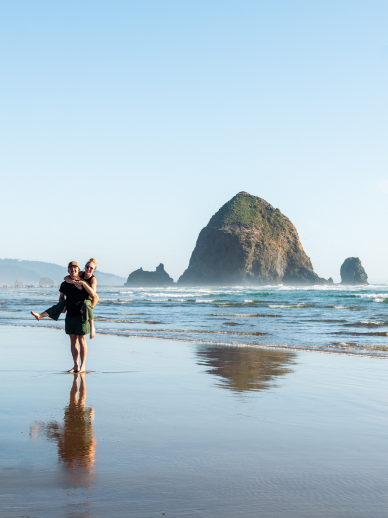 Couple posing playfully on the beach with Haystack Rock in the background at Cannon Beach, Oregon, during a sunny afternoon, a beloved coastal destination on a Pacific Northwest road trip.