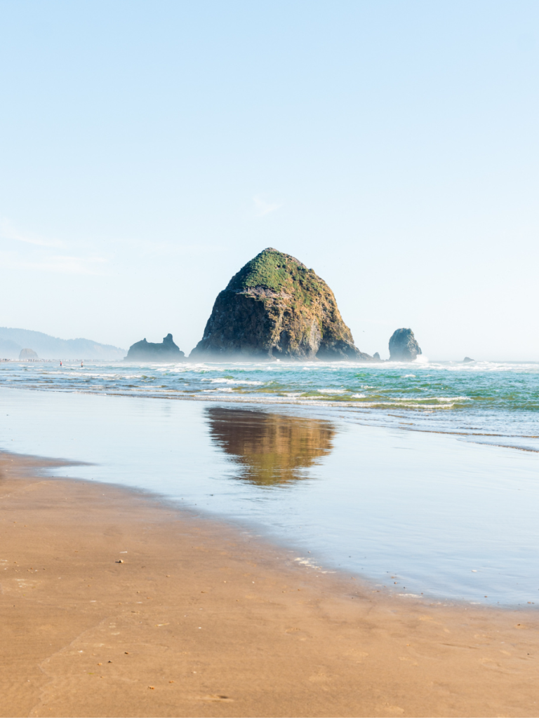 Haystack Rock reflected in the wet sand at low tide on Cannon Beach, Oregon, under a clear blue sky, a serene and iconic stop along the Pacific Northwest coast.