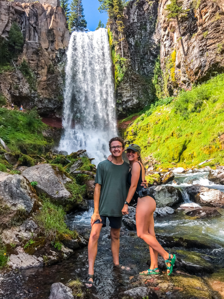 Couple standing in a shallow stream with a tall waterfall cascading behind them, surrounded by mossy cliffs and lush greenery, enjoying a refreshing stop on a Pacific Northwest road trip.