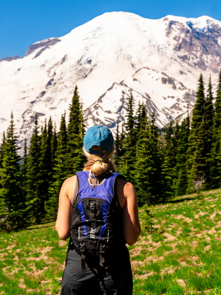 Woman wearing a backpack and blue cap stands facing Mount Rainier, taking in the snow-covered peak and surrounding evergreens on a sunny day, a classic hiking moment on a Pacific Northwest road trip.