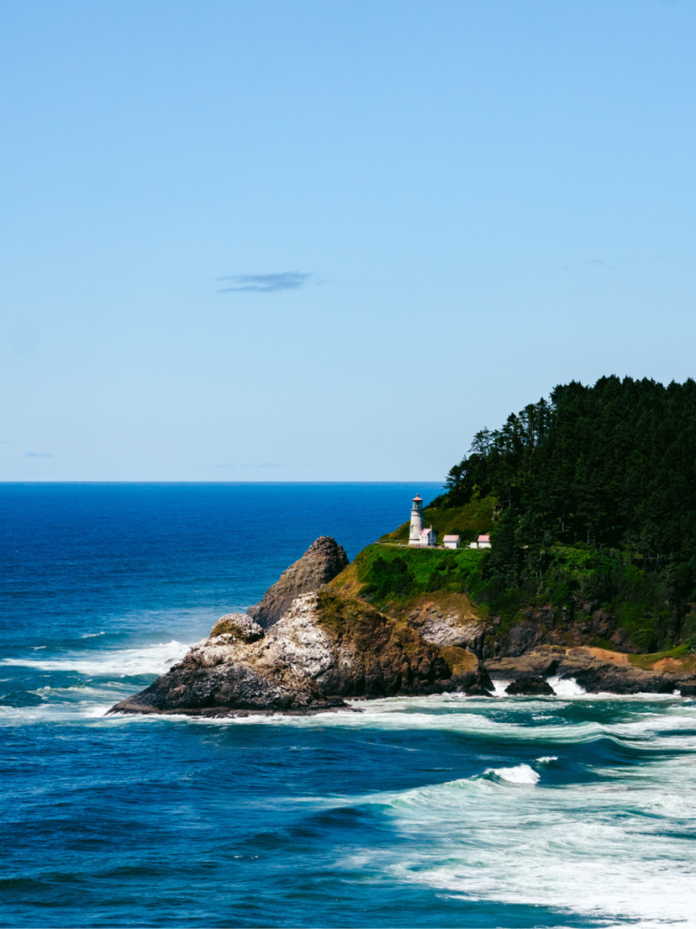 Heceta Head Lighthouse perched on a rugged cliff along the Oregon Coast, surrounded by evergreen trees and overlooking the deep blue Pacific Ocean with waves crashing below.