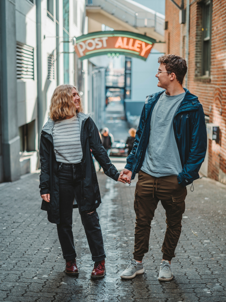 Couple holding hands and smiling at each other in front of the iconic Post Alley sign in Seattle, a charming stop on their Pacific Northwest road trip.