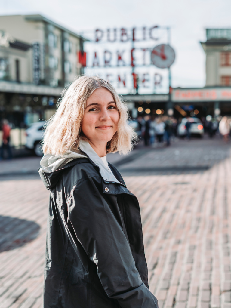 Young woman in a rain jacket smiling in front of Seattle’s iconic Pike Place Market sign, featured in a Seattle travel guide for first-time visitors.