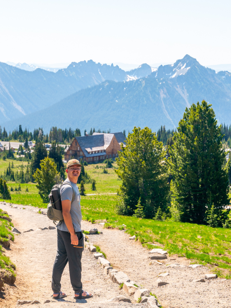 Man smiling on a mountain trail with a backpack, surrounded by lush greenery and rugged peaks near Paradise Lodge in Mount Rainier National Park, a scenic stop on a Pacific Northwest road trip.