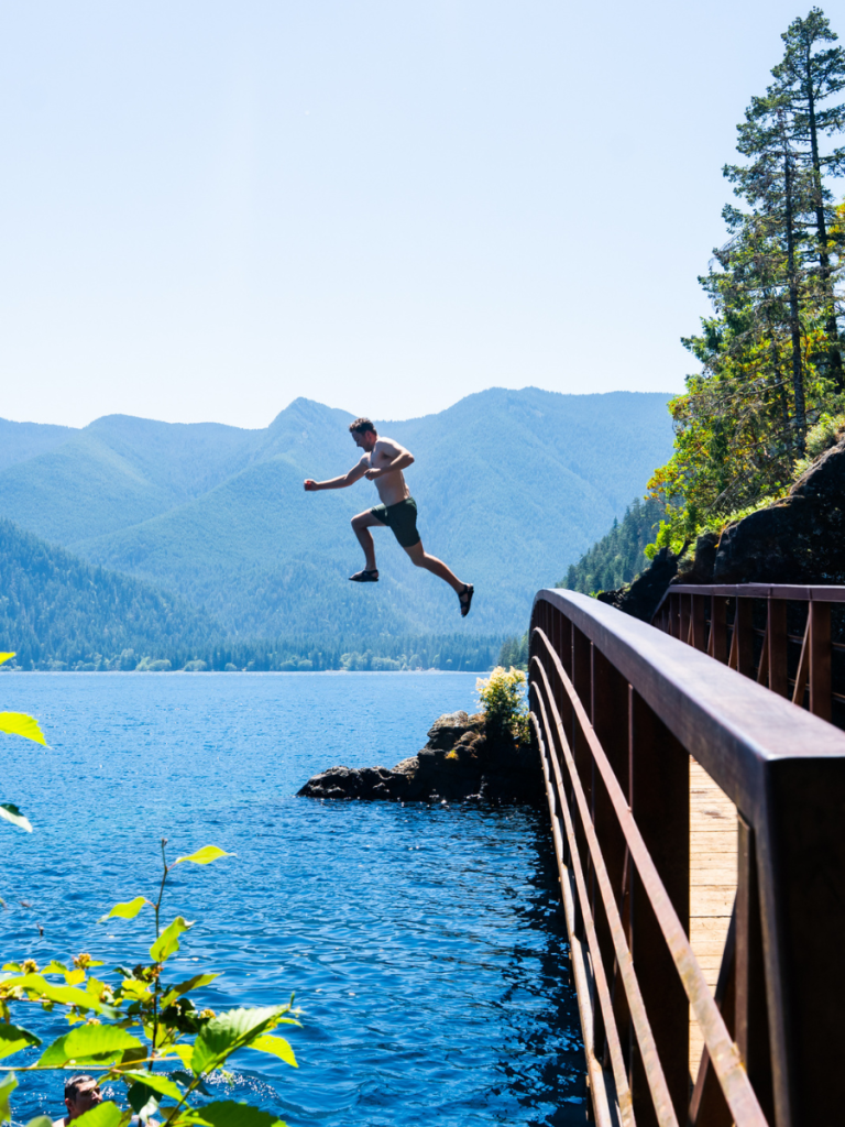 Man mid-air as he jumps off a bridge into the deep blue waters of Lake Crescent, with forested mountains in the background on a sunny summer day in Olympic National Park.