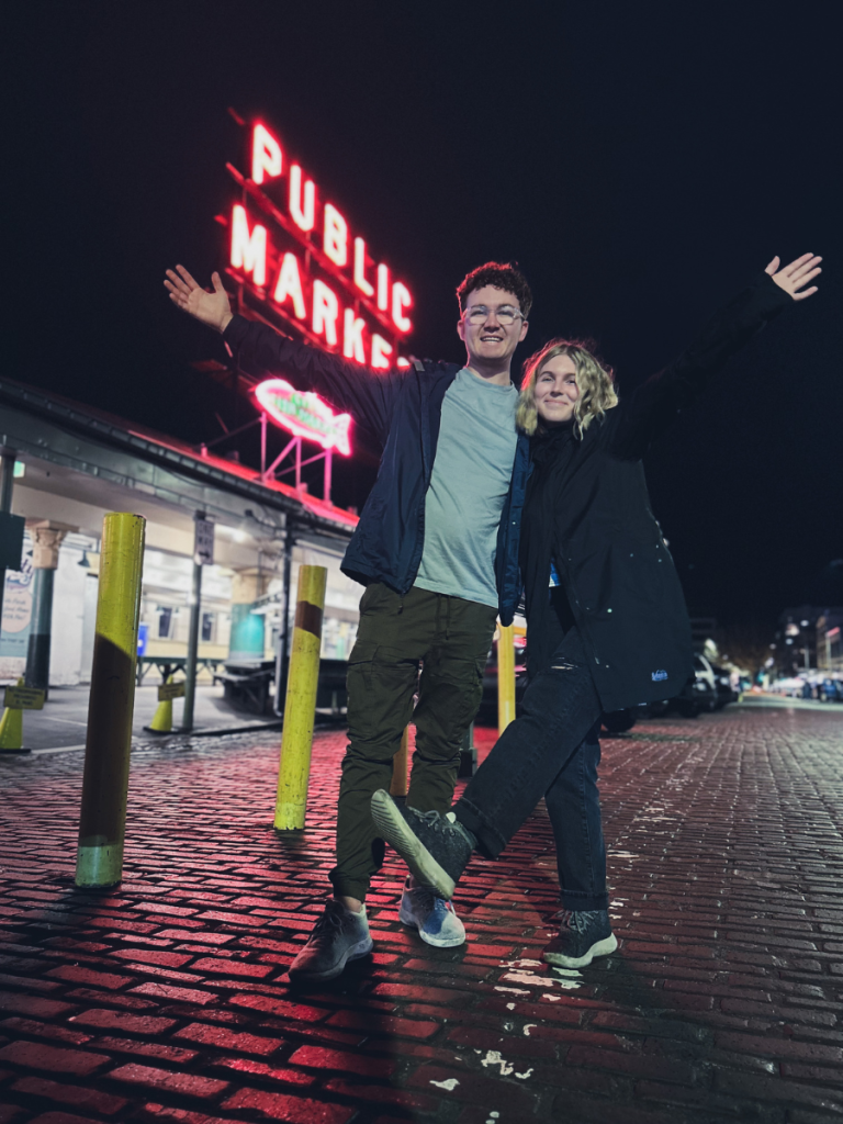 Smiling couple posing in front of the glowing Public Market sign at Seattle’s Pike Place Market during a nighttime stop on a Pacific Northwest road trip.