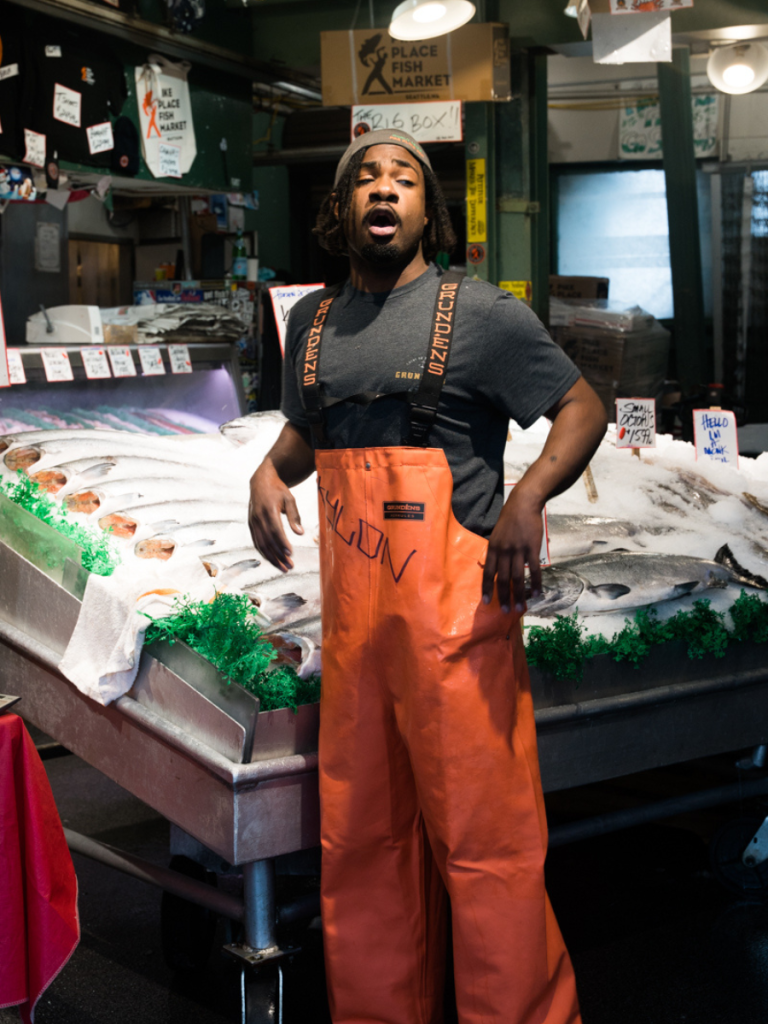 Fishmonger at Pike Place Fish Market in Seattle mid-shout, surrounded by fresh seafood on ice—an energetic and iconic stop on a Pacific Northwest road trip.