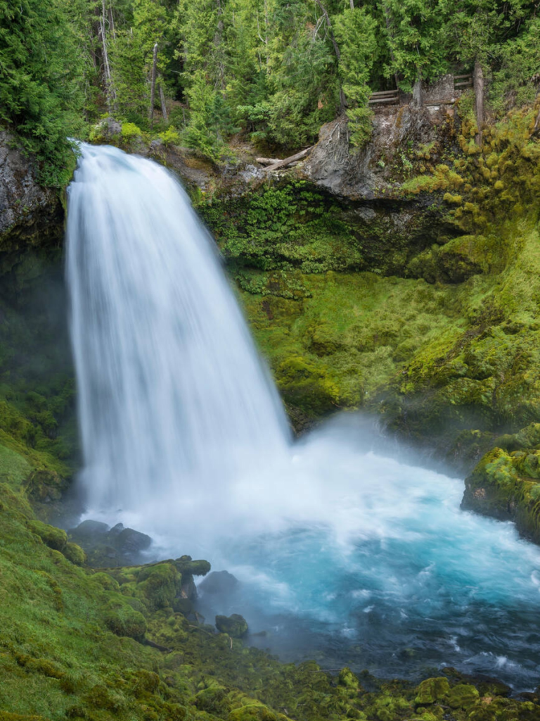 Crystal-clear waterfall plunging into a vibrant turquoise pool, surrounded by moss-covered rocks and dense evergreen forest, showcasing the lush beauty of the Pacific Northwest.