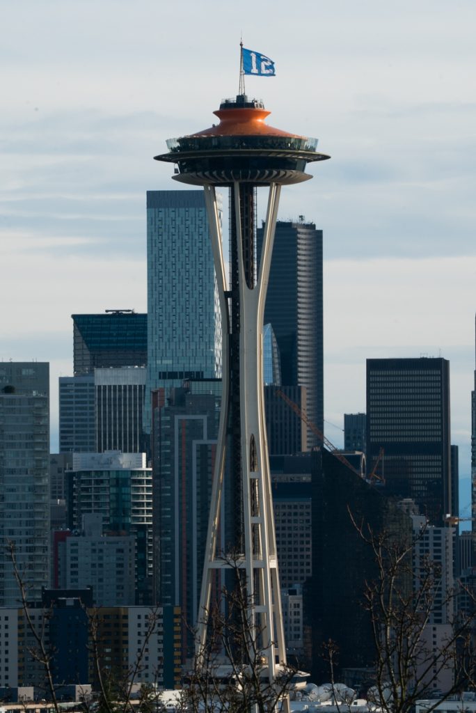 Close-up view of the Space Needle with a "12" Seahawks flag flying at the top, set against the Seattle skyline on a clear day, a must-see landmark on any Pacific Northwest road trip.
