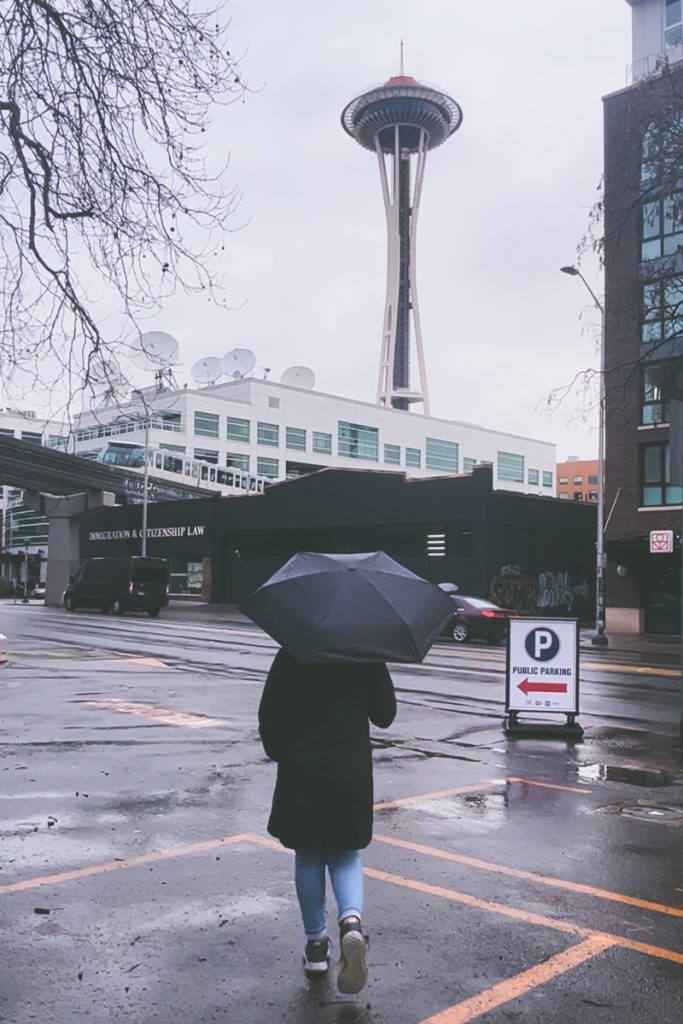 Person walking with a black umbrella on a rainy day in Seattle, with the Space Needle rising in the background—capturing classic Pacific Northwest weather on a road trip stop.