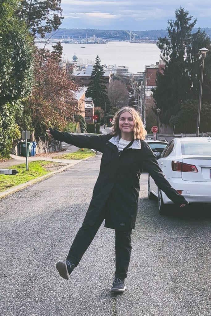 Smiling woman in a black raincoat posing playfully on a quiet street in Seattle, with a scenic view of Puget Sound and industrial cranes in the background, capturing a fun moment on a Pacific Northwest road trip.