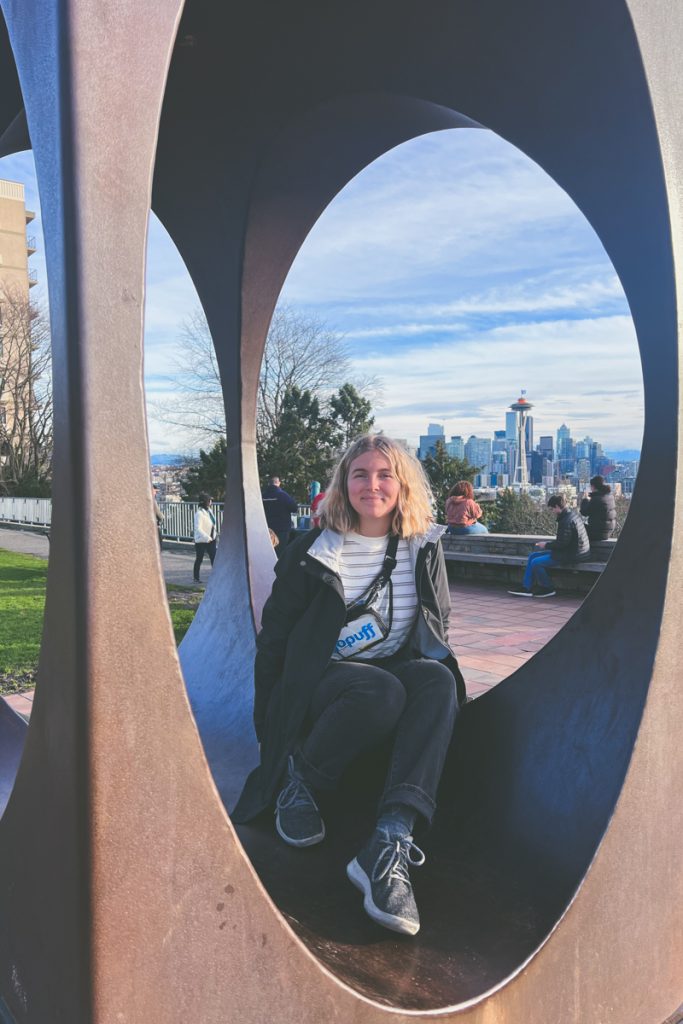 Woman sitting inside the viewpoint sculpture at Kerry Park in Seattle, with the city skyline and Space Needle framed perfectly in the background, a classic stop on a Pacific Northwest road trip.