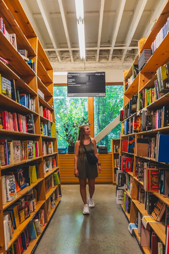 Woman browsing between tall, book-filled shelves inside Powell’s Books in Portland, Oregon, with large windows and greenery visible in the background.