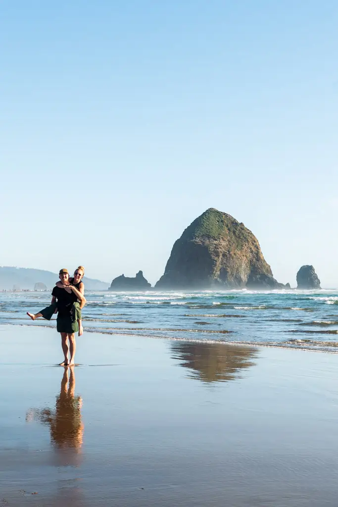 Two people standing on Cannon Beach with Haystack Rock towering in the background. Wet sand reflects the couple, while gentle waves roll onto the shore beneath a clear blue sky on the Oregon Coast.