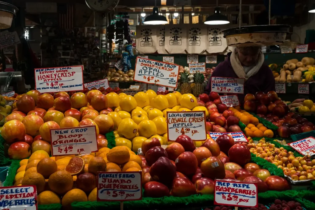 Colorful displays of fresh apples, oranges, berries, and other locally grown produce at Pike Place Market in Seattle, Washington, with handwritten vendor signs and bustling market stalls.