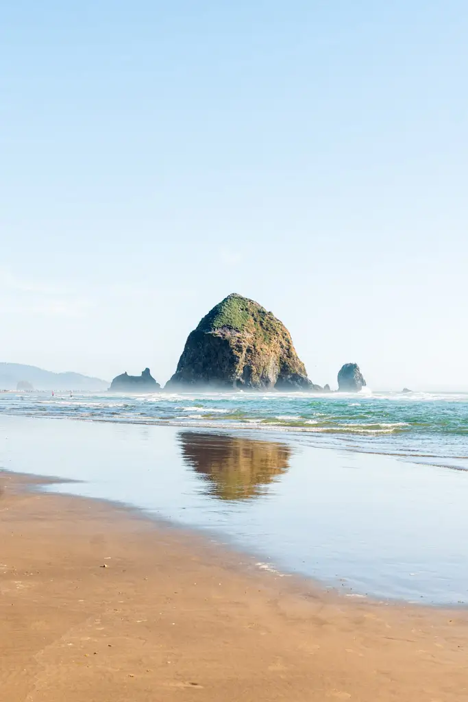 Haystack Rock rising from the Pacific Ocean at Cannon Beach, with gentle waves, a reflection on the wet sand, and clear blue skies along the iconic Oregon Coast.