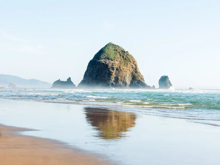 Haystack Rock rising from the Pacific Ocean at Cannon Beach, with gentle waves, a reflection on the wet sand, and clear blue skies along the Oregon Coast.