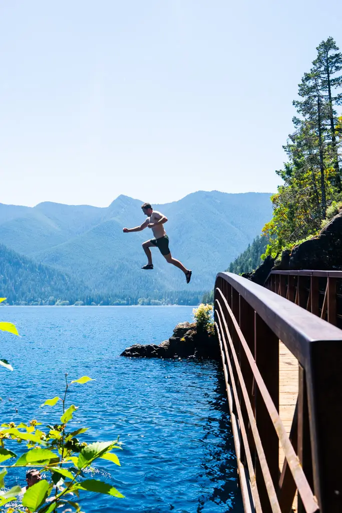 A person jumping from a bridge into the crystal-clear waters of Lake Crescent, surrounded by forested mountains and scenic views in Olympic National Park, Washington.