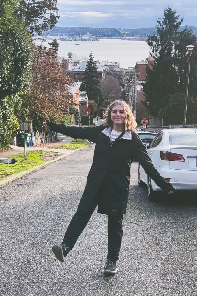 A person standing in the middle of a residential street near Kerry Park with Elliott Bay and the Seattle skyline visible in the distance on a cloudy day.