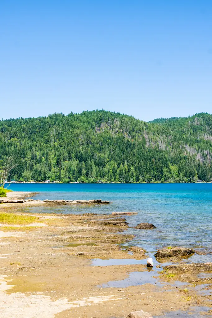 The turquoise shoreline of Lake Crescent with calm, crystal-clear water, a forested mountain backdrop, and a peaceful lakeside beach in Olympic National Park, Washington.