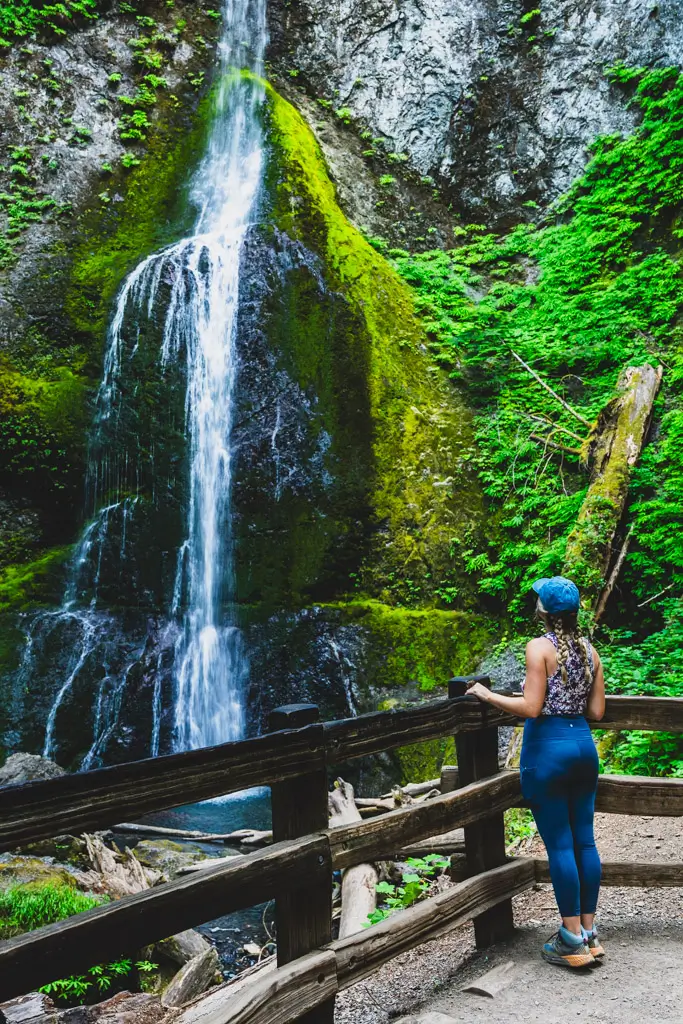 A traveler stands at the viewing platform overlooking Marymere Falls as the waterfall cascades down a moss-covered cliff surrounded by lush green forest in Olympic National Park, Washington.