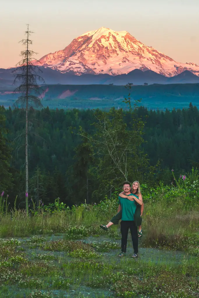 A couple posing with Mount Rainier glowing at sunset in the background, surrounded by wildflowers and evergreen forests at a scenic viewpoint in Mount Rainier National Park, Washington.