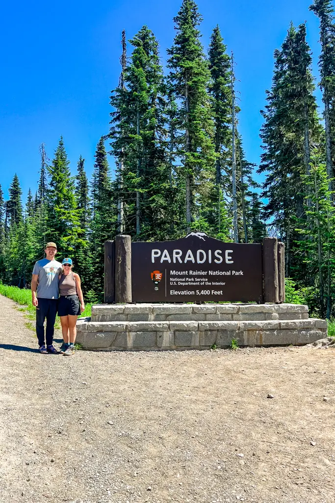 A couple standing beside the Paradise entrance sign at 5,400 feet in Mount Rainier National Park, Washington, surrounded by towering evergreen trees on a sunny summer day.