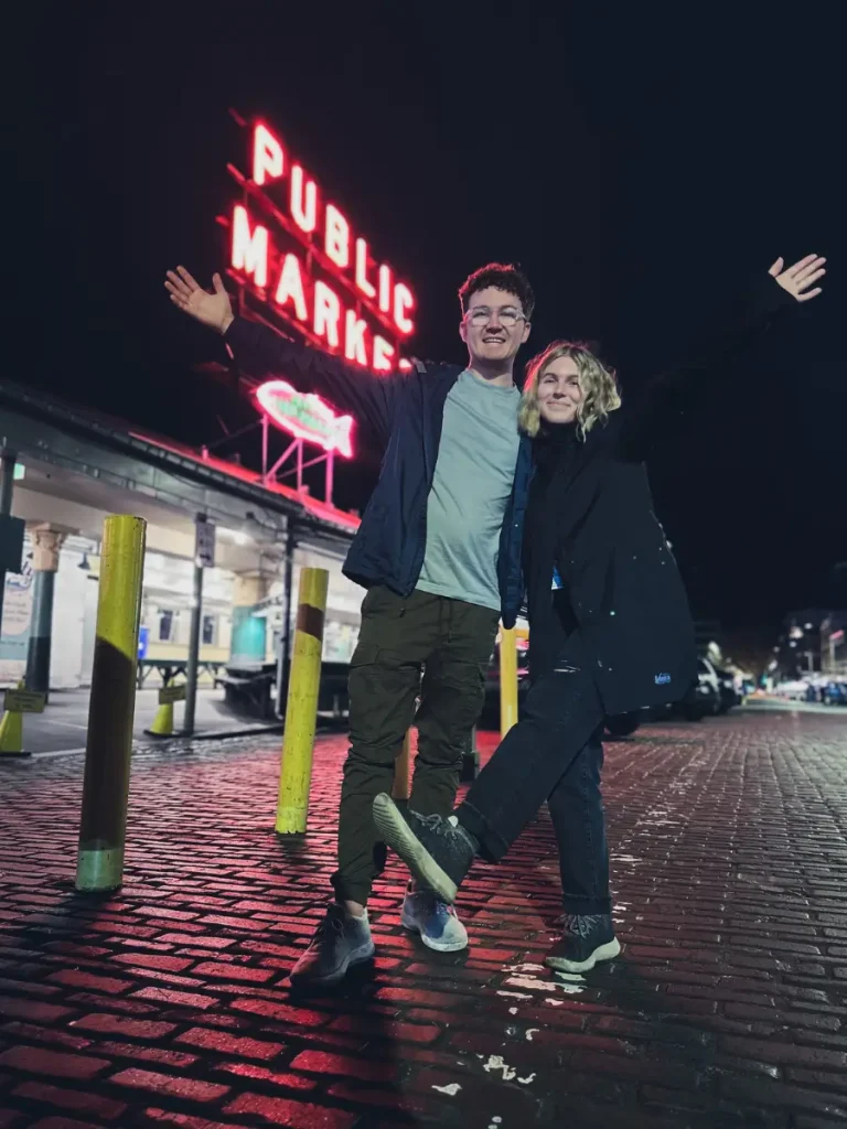 A couple posing beneath the glowing Pike Place Market sign at night, with the famous neon landmark and wet cobblestone streets in downtown Seattle, Washington.