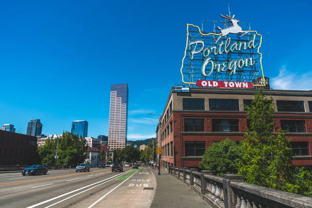 Iconic Portland Oregon neon sign above a historic Old Town building, overlooking a downtown street with the Portland skyline under a bright blue sky.