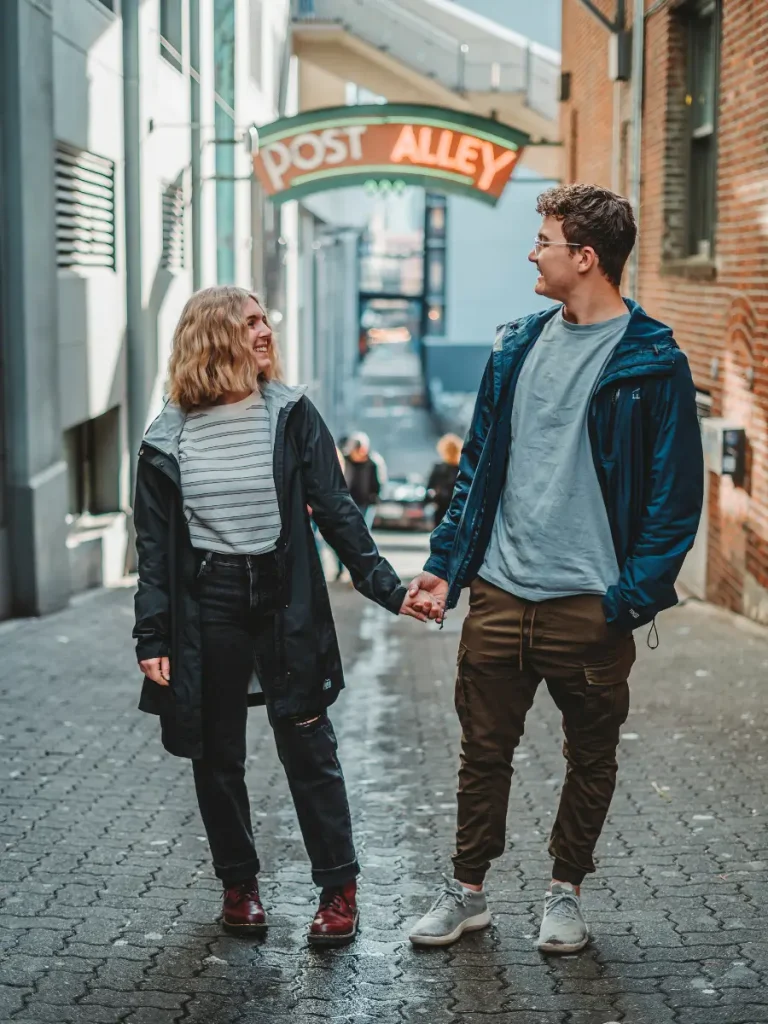 A couple holding hands while walking through Seattle's Post Alley, with the iconic Post Alley sign overhead and the historic streets of downtown Seattle in the background.
