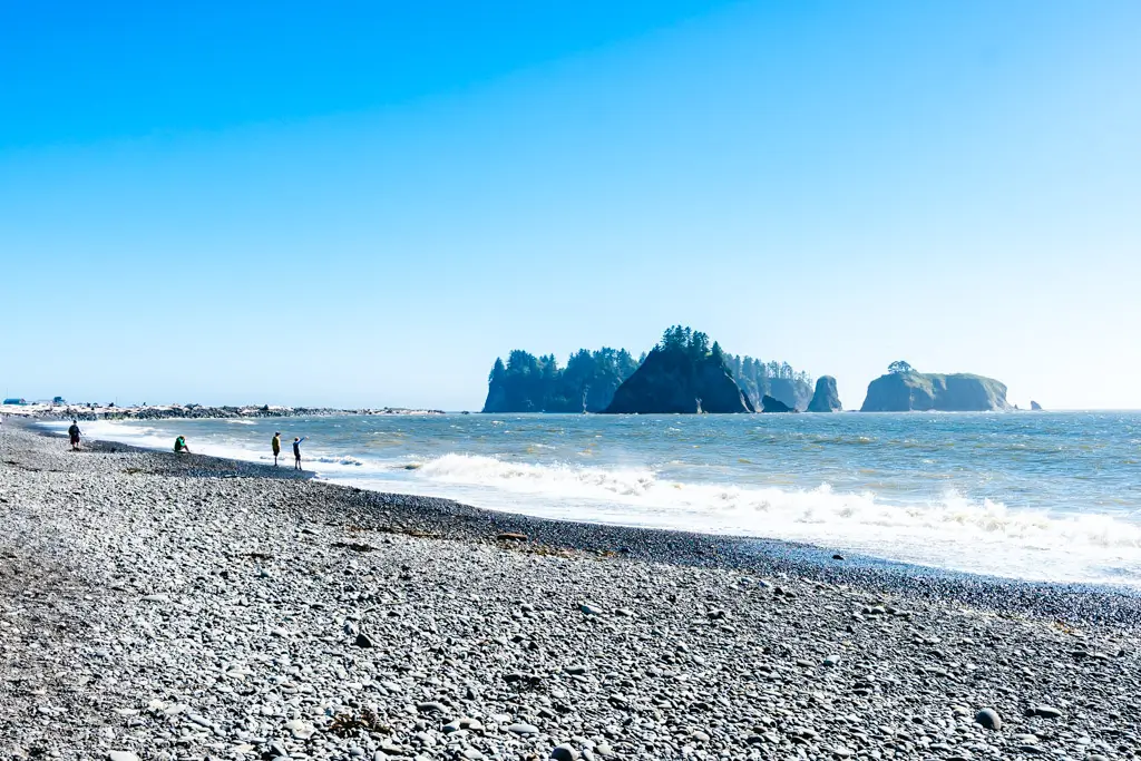 Rialto Beach in Olympic National Park with a rocky shoreline, crashing Pacific waves, and dramatic sea stacks rising from the ocean beneath a bright blue sky in Washington.