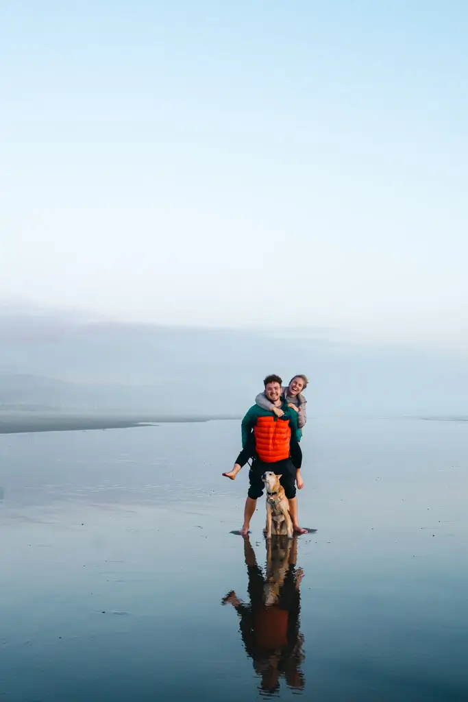A couple with their dog standing on a wide, reflective beach at sunset along the Oregon Coast, with calm water, soft pastel skies, and their reflections mirrored in the wet sand.
