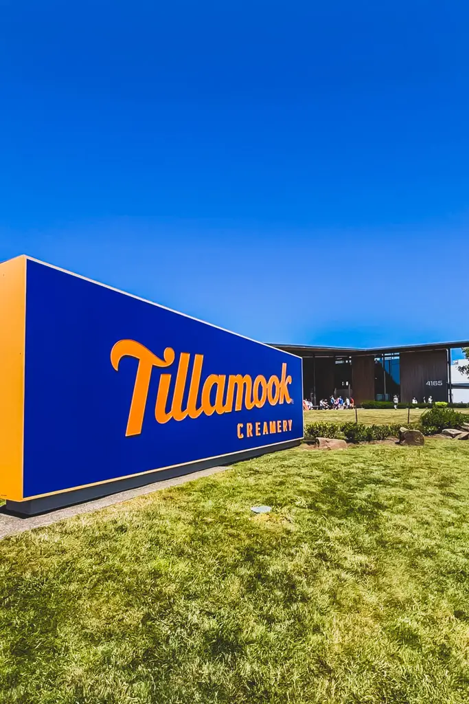 The iconic Tillamook Creamery sign outside the visitor center in Tillamook, Oregon, with the modern creamery building and green lawn under a bright blue sky.