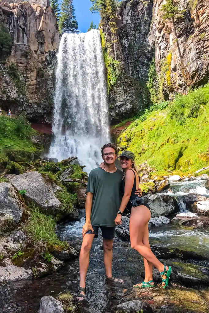 Couple standing in a shallow stream with a tall waterfall cascading behind them, surrounded by mossy cliffs and lush greenery, enjoying a refreshing stop on a Pacific Northwest road trip.