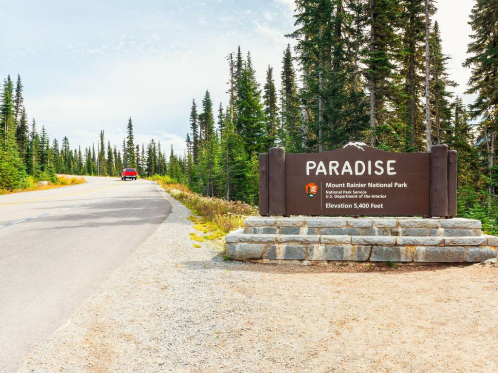 Wooden entrance sign for Paradise in Mount Rainier National Park, with a red car driving up a forested mountain road.