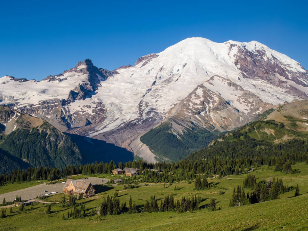 Snow-covered Mount Rainier towering above green alpine meadows at Sunrise Mount Rainier National Park, with the visitor center and surrounding forest in view under a clear blue sky.