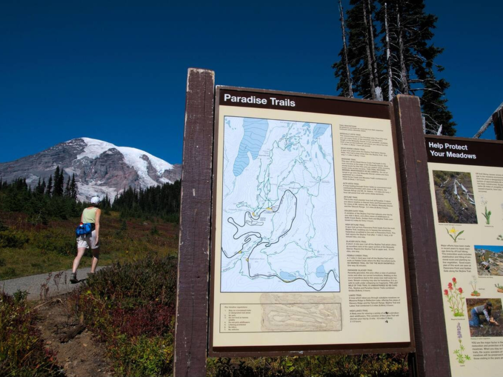 Hiker walking past the Paradise Trails sign in Mount Rainier National Park Paradise with the snow-capped peak rising in the background.