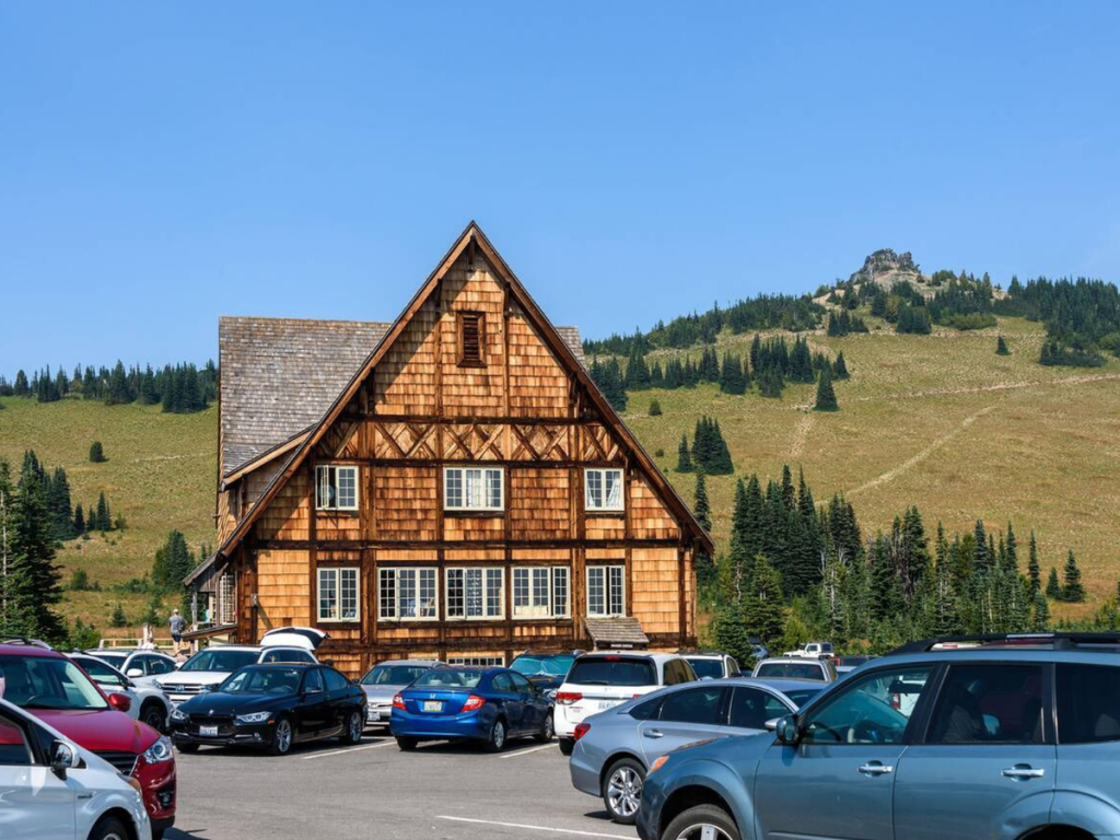 Wooden Sunrise Visitor Center at Mount Rainier National Park, surrounded by parked cars with rolling green hills and evergreen trees in the background.