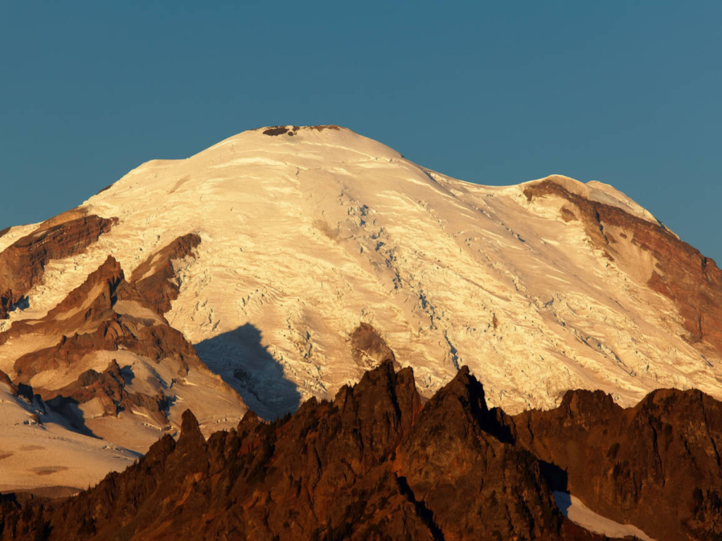 Snow-covered summit of Mount Rainier glowing golden in the early morning light at Sunrise Mount Rainier National Park, with jagged dark peaks in the foreground.
