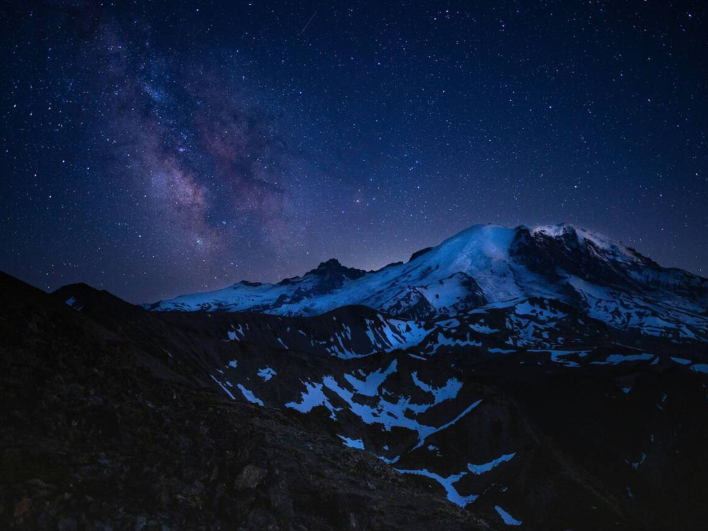 Star-filled night sky with the Milky Way arching above the snow-covered peaks of Mount Rainier at Sunrise Mount Rainier National Park.