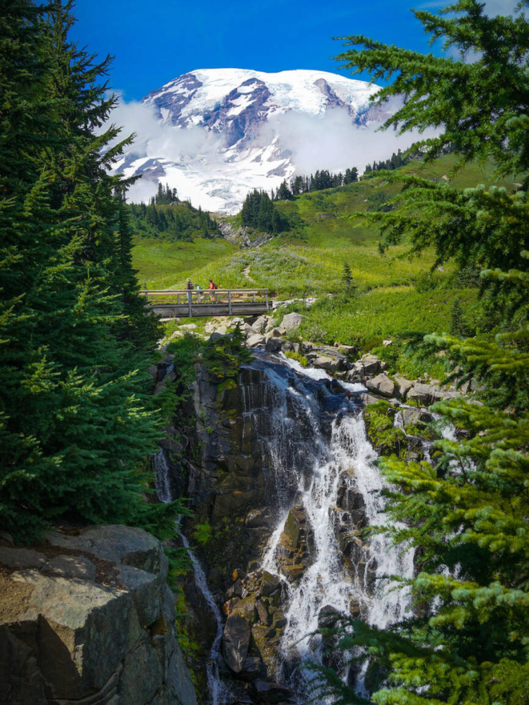 Scenic waterfall cascading beneath a wooden footbridge with hikers, framed by evergreens and backed by snow-covered Mount Rainier on a sunny day.