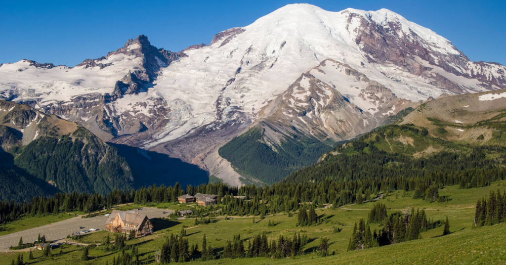 Snow-covered Mount Rainier towering above green alpine meadows at Sunrise Mount Rainier National Park, with the visitor center and surrounding forest in view under a clear blue sky.