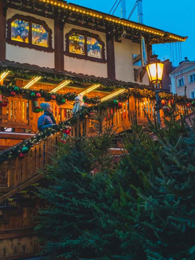 Two people stand on a warmly lit wooden staircase lined with garlands and ornaments, outside a cozy chalet-style building nestled among fresh pine trees — a festive and intimate moment at a European Christmas market.