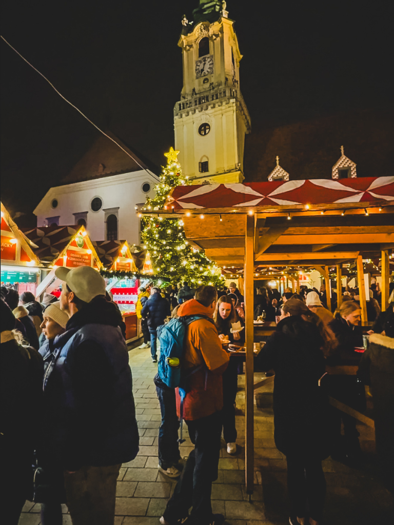 Visitors gather under festive lights and a Christmas tree at the Christmas markets in Bratislava, with the historic Old Town Hall tower glowing in the background.