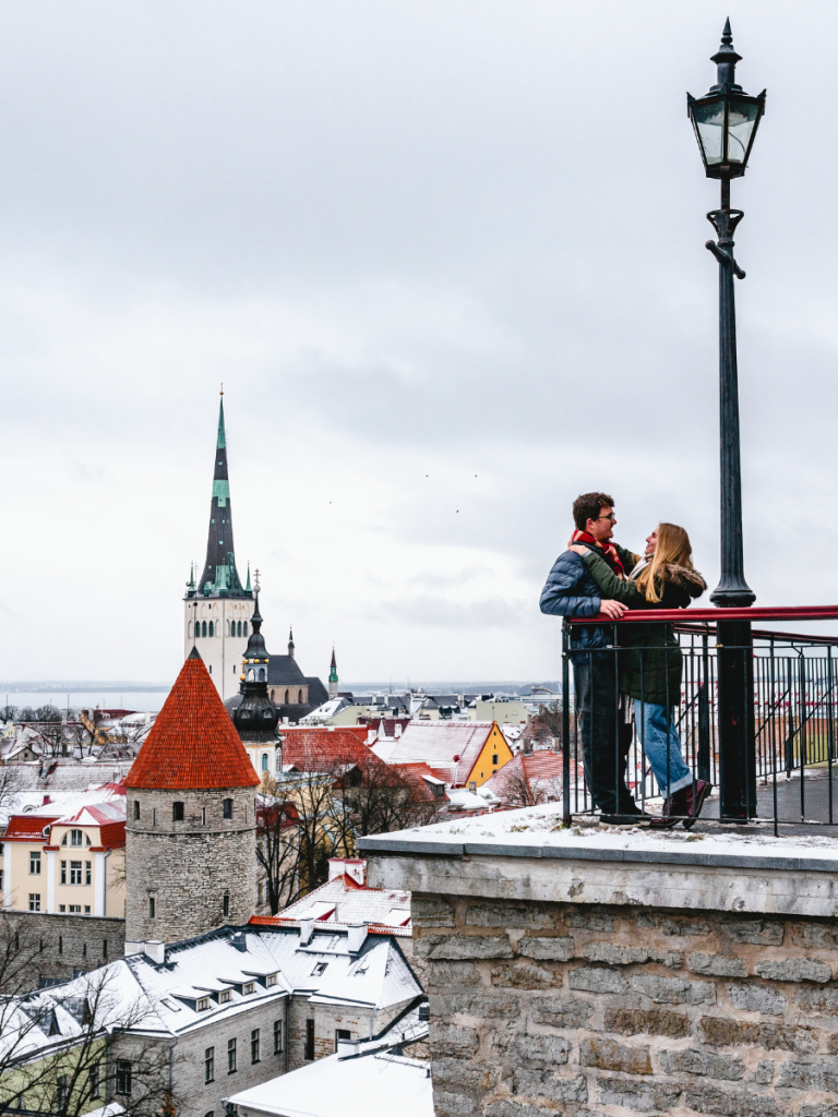 A couple embraces at a snowy viewpoint overlooking Tallinn’s Old Town rooftops and church towers — winter romance and scenic views during European Christmas markets season.